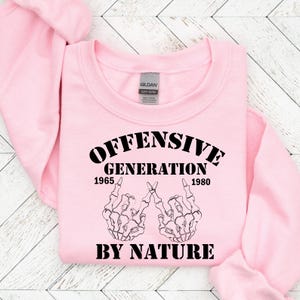 May include: Pink sweatshirt with black text that reads "Offensive Generation 1965 - 1980 By Nature". The text is surrounded by two skeleton hands making the middle finger gesture.