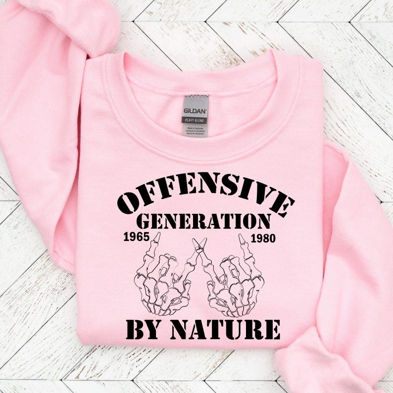 May include: Pink sweatshirt with black text that reads "Offensive Generation 1965 - 1980 By Nature". The text is surrounded by two skeleton hands making the middle finger gesture.