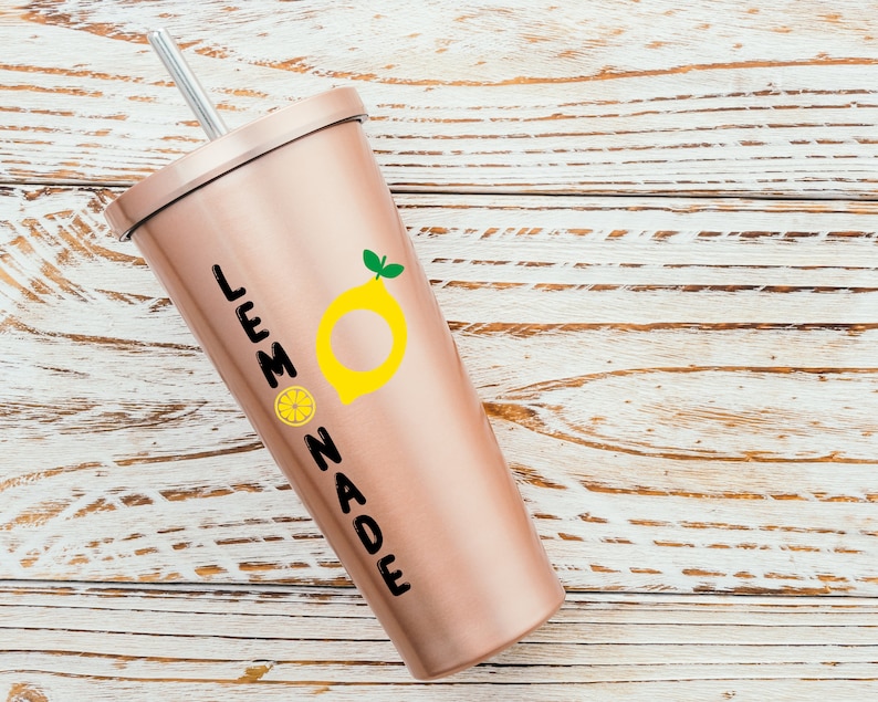 Puede incluir: Un vaso de color rosa dorado con un gr&aacute;fico de lim&oacute;n negro y amarillo y el texto "LEMONADE".