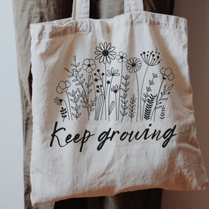Puede incluir: Una bolsa de tela blanca con dibujos de flores en negro y el texto "Keep growing".