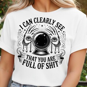以下が含まれることがあります： 黒いグラフィックの白いTシャツ。デザインは、目の付いた2つの手の間にクリスタルボールがあり、「I CAN CLEARLY SEE THAT YOU ARE FULL OF SHIT」というテキストが特徴です。デザインは神秘的なテーマです。