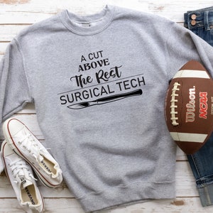 May include: A gray sweatshirt with the text "A cut above the rest surgical tech" and a black illustration of a scalpel. A football and a pair of white sneakers are also in the image.