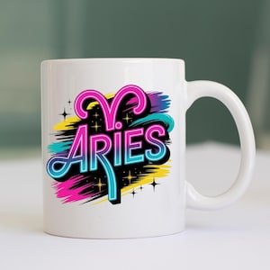 Op de afbeelding: Witte keramische mok met het woord "ARIES" in een kleurrijk, gestileerd ontwerp. Het ontwerp bevat roze, blauwe, gele en zwarte elementen, met steraccenten. De mok heeft een gebogen handvat.
