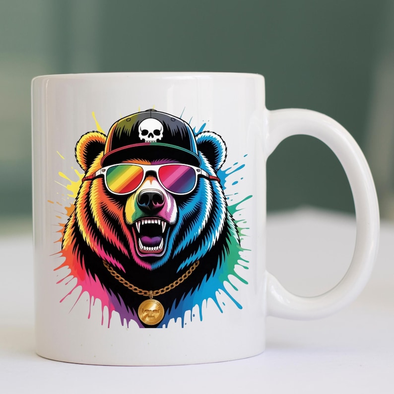 May include: White ceramic mug featuring a colorful illustration of a bear wearing a black cap with a skull, rainbow sunglasses, and a gold chain. The bear has an open mouth and is surrounded by colorful paint splatters.