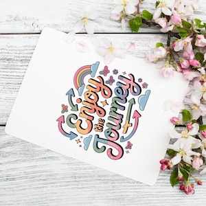 Op de afbeelding: Een witte kaart met de tekst "Enjoy the Journey" in kleurrijke, speelse letters. Het ontwerp bevat een regenboog, vlinders, sterren en pijlen. De kaart wordt omringd door witte en roze bloemen op een lichte houten achtergrond.