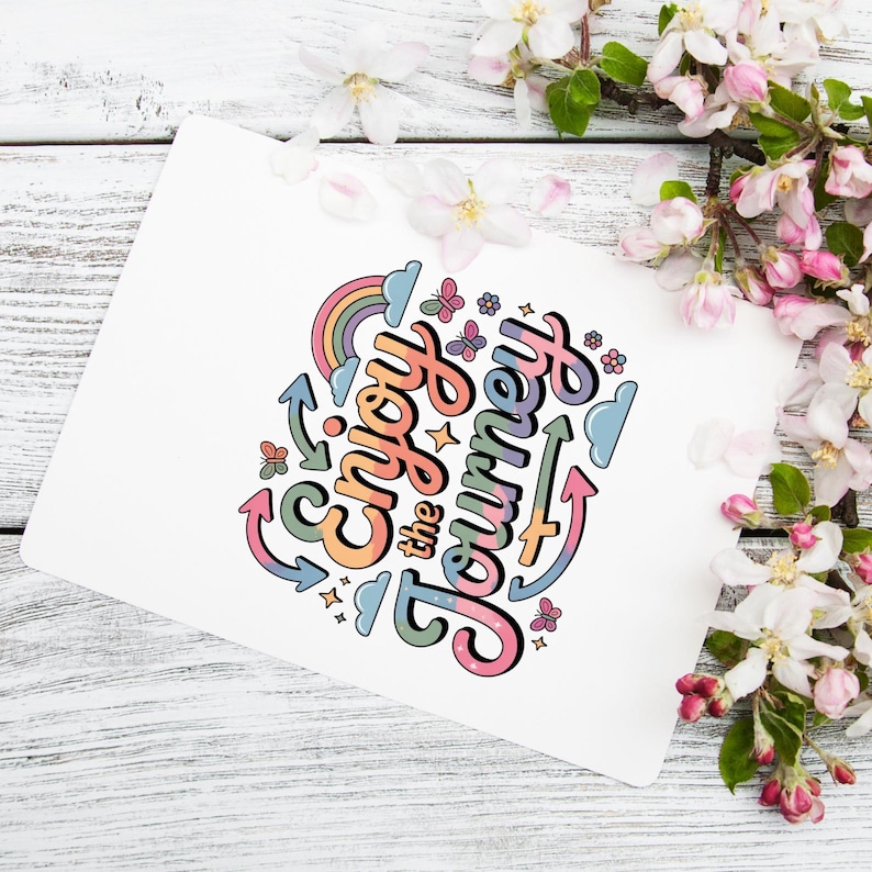 Op de afbeelding: Een witte kaart met de tekst "Enjoy the Journey" in kleurrijke, speelse letters. Het ontwerp bevat een regenboog, vlinders, sterren en pijlen. De kaart wordt omringd door witte en roze bloemen op een lichte houten achtergrond.