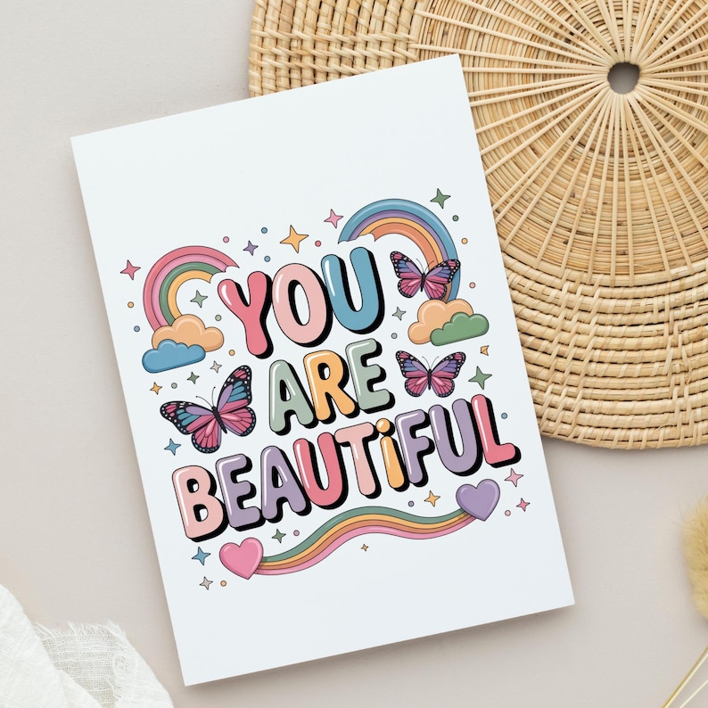 Op de afbeelding: Een witte kaart met de tekst "YOU ARE BEAUTIFUL" in kleurrijke letters. Het ontwerp bevat regenbogen, vlinders, wolken, sterren en harten. De kaart ligt op een licht oppervlak, met een gevlochten mand op de achtergrond.