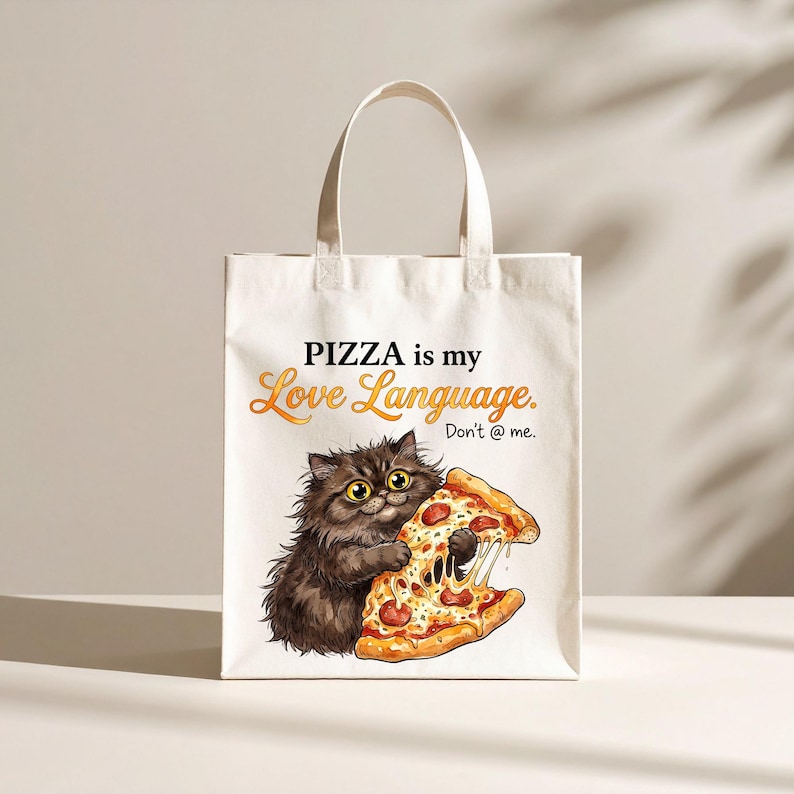 May include: A white tote bag featuring a graphic of a fluffy dark brown cat holding a slice of pepperoni pizza. The text reads PIZZA is my Love Language. Don't @ me. The bag has two top handles and a rectangular shape with a clean, flat design.