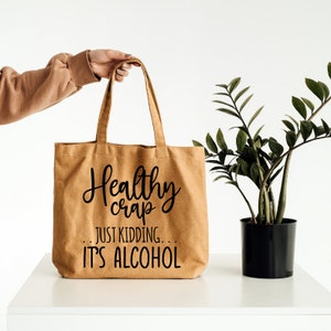May include: A brown canvas tote bag with black text that reads "Healthy crap... Just kidding... It's alcohol".