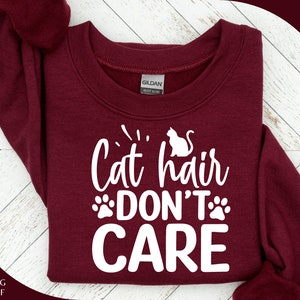 Może przedstawiać: Bordowy sweter z białym napisem "Cat hair don't care" z odciskiem łapy po obu stronach słowa "don't".