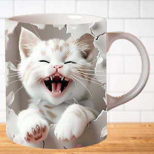 Pu&ograve; includere: Tazza in ceramica bianca con un'immagine 3D di un gattino bianco con macchie marroni che spunta da un buco nella tazza. Il gattino ha la bocca aperta e ride.