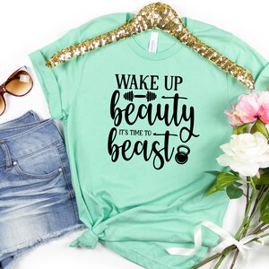 Op de afbeelding: Een mintgroen T-shirt met de tekst "Wake Up Beauty It's Time To Beast" in zwarte letters. De tekst bevat een halter en een kettlebell.