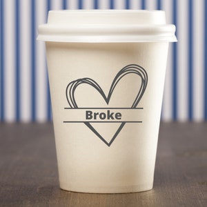 May include: White to-go coffee cup with a black heart and the word "Broke" written on a banner across the heart.