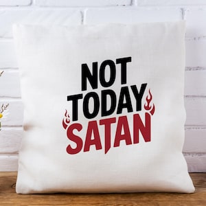 K&ouml;nnte beinhalten: Ein wei&szlig;es quadratisches Kissen mit dem Aufdruck "NOT TODAY SATAN" in Schwarz und Rot. Flammen befinden sich auf beiden Seiten des Wortes "SATAN". Das Kissen befindet sich vor einer wei&szlig;en Backsteinmauer.