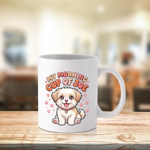 May include: A white ceramic mug featuring a colorful cartoon illustration of a small, fluffy, light brown and white puppy with a pink collar. Above the puppy, the text reads "MY MORNING CUP OF JOE" in orange letters, surrounded by small pink paw prints.