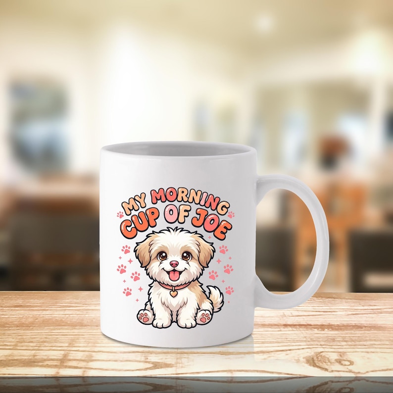 May include: A white ceramic mug featuring a colorful cartoon illustration of a small, fluffy, light brown and white puppy with a pink collar. Above the puppy, the text reads "MY MORNING CUP OF JOE" in orange letters, surrounded by small pink paw prints.