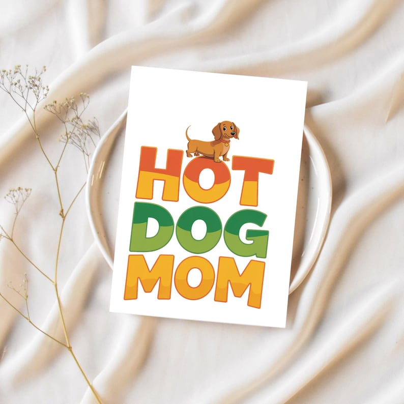 May include: A white card with the words "HOT DOG MOM" in orange, green, and yellow block letters. A cartoon dachshund dog is perched above the word "HOT". The card is on a white plate with a sprig of dried flowers.