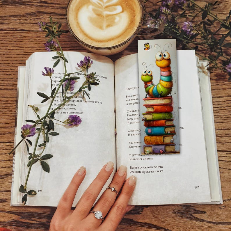 May include: An open book with text, a colorful caterpillar bookmark, a cup of coffee, and purple flowers on a wooden surface. The bookmark features a stack of books with a cartoon caterpillar design.