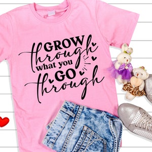 May include: Pink t-shirt with black text that reads "GROW through what you GO through" with small hearts around the text. The shirt is lying on a white wooden surface with a pair of blue jeans, two red hearts, and two stuffed toys.