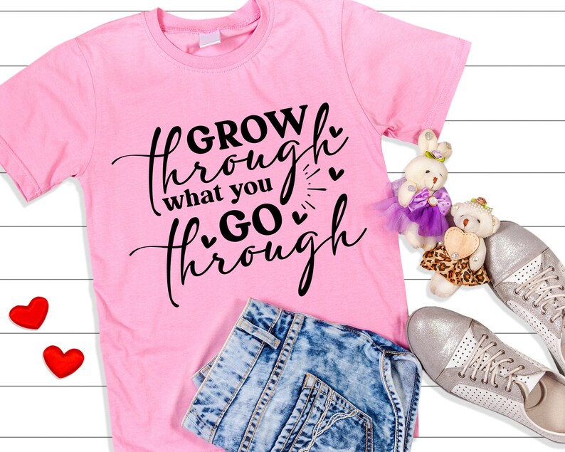 May include: Pink t-shirt with black text that reads "GROW through what you GO through" with small hearts around the text. The shirt is lying on a white wooden surface with a pair of blue jeans, two red hearts, and two stuffed toys.