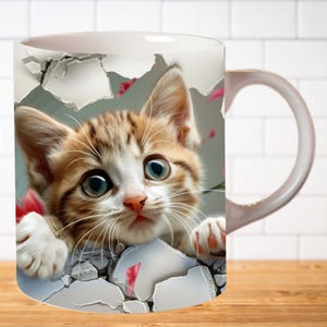 Pu&ograve; includere: Tazza in ceramica bianca con un'immagine 3D di un gattino marrone e bianco che spunta da un muro screpolato. Il gattino ha gli occhi azzurri e dettagli rosa.