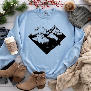 May include: A light blue sweatshirt with a black graphic of mountains and trees in a diamond shape.