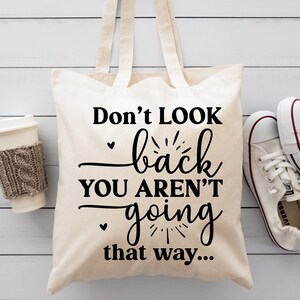 May include: A white canvas tote bag with black text that reads "Don't look back you aren't going that way..." The bag is sitting on a white wooden surface with a cup of coffee and a pair of white sneakers.