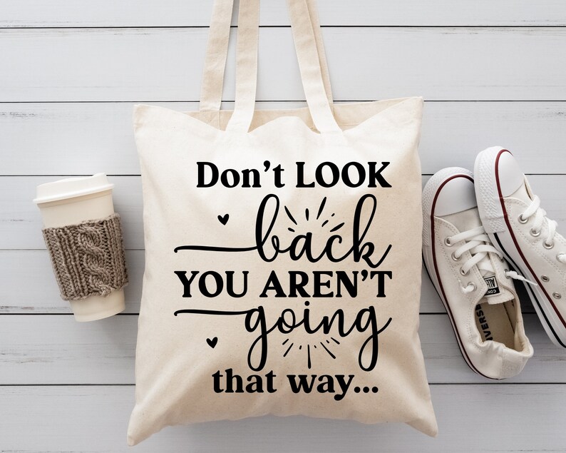 May include: A white canvas tote bag with black text that reads "Don't look back you aren't going that way..." The bag is sitting on a white wooden surface with a cup of coffee and a pair of white sneakers.