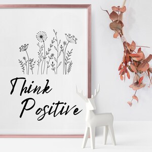 May include: A framed print with a black and white illustration of wildflowers and the text "Think Positive". A white ceramic deer figurine is in the foreground.