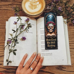 May include: An open book with a sprig of purple flowers, a coffee cup, and a bookmark. The bookmark features a skull with a floral crown and the text "Dead Tired But Still Reading."