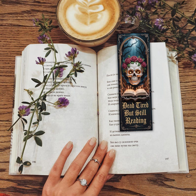 May include: An open book with a sprig of purple flowers, a coffee cup, and a bookmark. The bookmark features a skull with a floral crown and the text "Dead Tired But Still Reading."