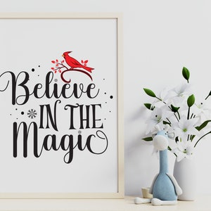May include: A white framed print with a black and white cursive font that reads "Believe in the Magic". A red cardinal bird is perched on a branch with green leaves. The background is a white wall.