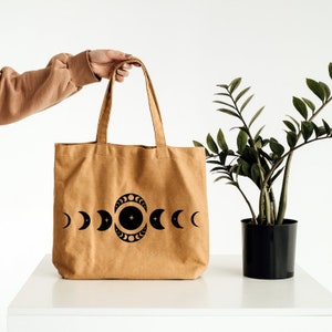 May include: A tan canvas tote bag with a black celestial design featuring phases of the moon and stars. The bag has two handles and is held up by a person. A potted plant is to the right.