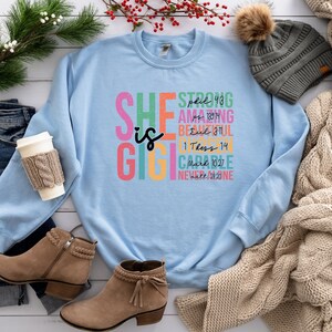 K&ouml;nnte beinhalten: Hellblauer Pullover mit dem Text "SHE is GIGIBLE" in bunten Buchstaben. Der Text ist vertikal gestapelt mit Bibelversen und Daten neben jedem Wort.