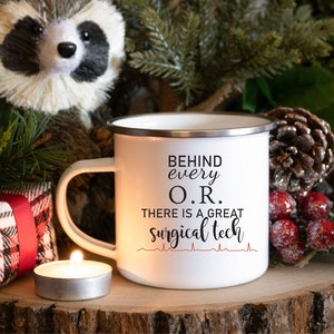 May include: White enamel mug with a black and red design. The mug says "Behind every O.R. there is a great surgical tech" with a red heartbeat graphic.