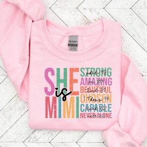 K&ouml;nnte beinhalten: Rosafarbener Pullover mit dem Text "SHE is MIGHTY" in Regenbogenfarben, unter jedem Wort sind Bibelverse aufgelistet.
