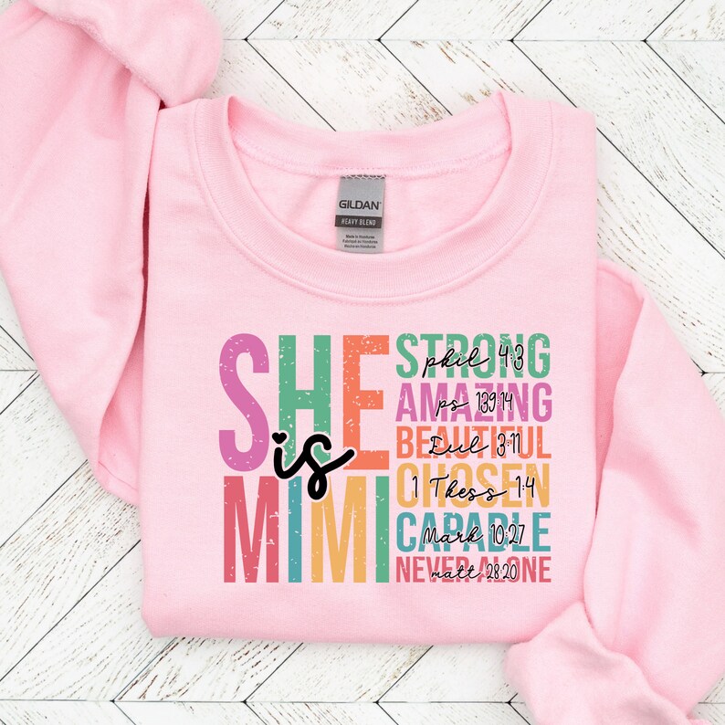 K&ouml;nnte beinhalten: Rosafarbener Pullover mit dem Text "SHE is MIGHTY" in Regenbogenfarben, unter jedem Wort sind Bibelverse aufgelistet.