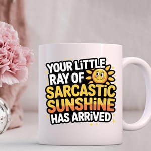 K&ouml;nnte beinhalten: Wei&szlig;e Keramik-Tasse mit dem Text "YOUR LITTLE RAY OF SARCASTIC SUNSHINE HAS ARRIVED" in Schwarz, Gelb und Orange. Das Design beinhaltet eine l&auml;chelnde Sonne. Die Tasse hat einen gebogenen Henkel.