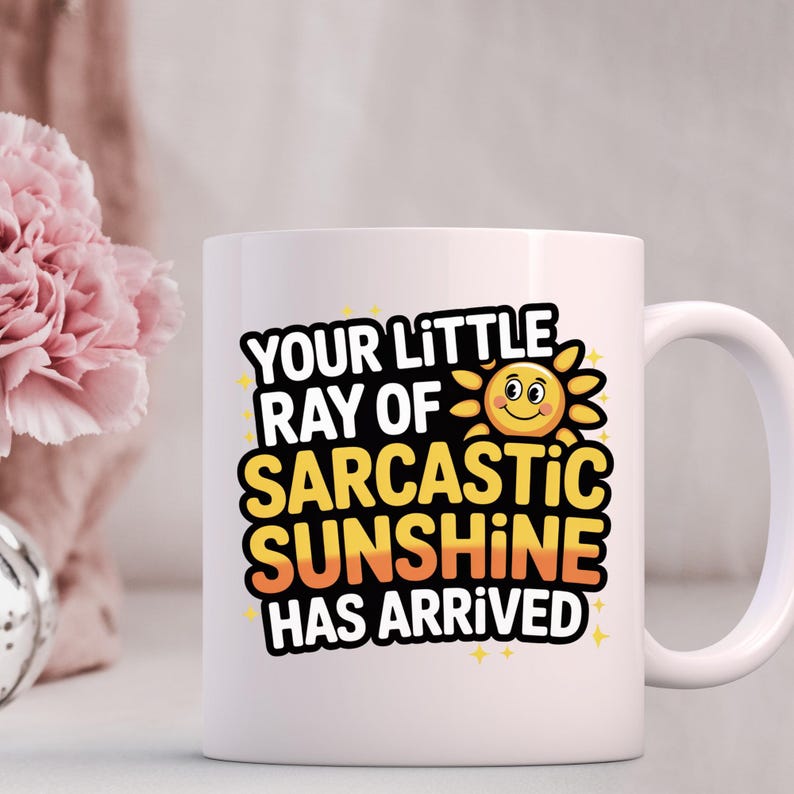K&ouml;nnte beinhalten: Wei&szlig;e Keramik-Tasse mit dem Text "YOUR LITTLE RAY OF SARCASTIC SUNSHINE HAS ARRIVED" in Schwarz, Gelb und Orange. Das Design beinhaltet eine l&auml;chelnde Sonne. Die Tasse hat einen gebogenen Henkel.