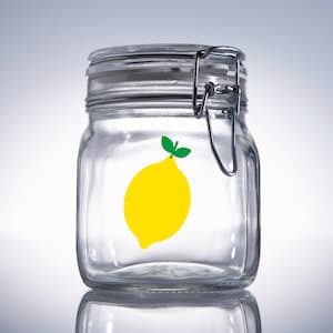 Puede incluir: Un tarro de vidrio transparente con tapa de metal y una pegatina de lim&oacute;n amarillo en la parte delantera. El tarro est&aacute; vac&iacute;o.