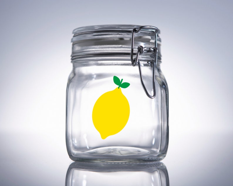 Puede incluir: Un tarro de vidrio transparente con tapa de metal y una pegatina de lim&oacute;n amarillo en la parte delantera. El tarro est&aacute; vac&iacute;o.