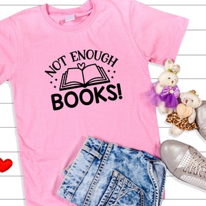 May include: Pink t-shirt with the text "Not Enough Books!" and a black and white illustration of an open book with a heart on the cover. The shirt is laid out on a white wooden surface with a pair of blue jeans, two red hearts, and two stuffed animals.