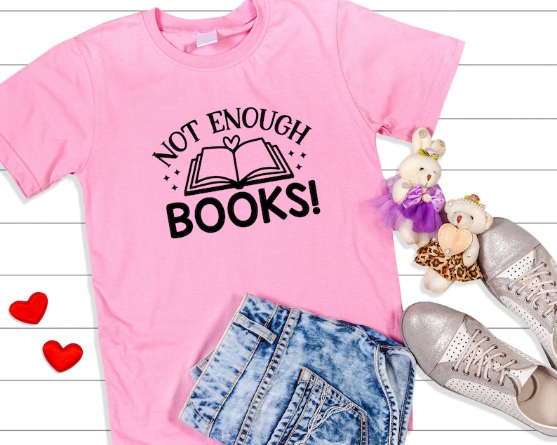 May include: Pink t-shirt with the text "Not Enough Books!" and a black and white illustration of an open book with a heart on the cover. The shirt is laid out on a white wooden surface with a pair of blue jeans, two red hearts, and two stuffed animals.