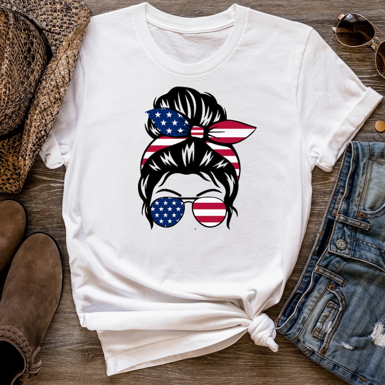 K&ouml;nnte beinhalten: Wei&szlig;es T-Shirt mit einem Motiv einer Frau mit einem unordentlichen Dutt, die eine Sonnenbrille mit amerikanischem Flaggenmuster tr&auml;gt. Der Dutt ist auch mit einem rot-wei&szlig;-blauen Band verziert.
