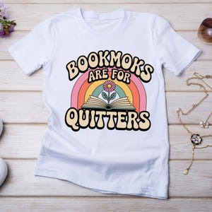 以下が含まれることがあります： 虹、開いた本と花、そして「BOOKMOKS ARE FOR QUITTERS」というテキストが遊び心のあるフォントでデザインされた、レトロなデザインの白いTシャツ。 このシャツは柔らかい素材でできており、カジュアルウェアに適しています。