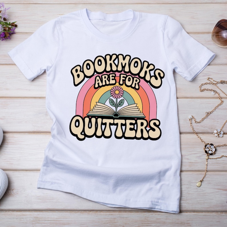 以下が含まれることがあります： 虹、開いた本と花、そして「BOOKMOKS ARE FOR QUITTERS」というテキストが遊び心のあるフォントでデザインされた、レトロなデザインの白いTシャツ。 このシャツは柔らかい素材でできており、カジュアルウェアに適しています。