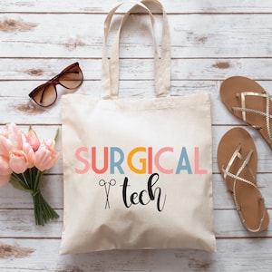 May include: A white canvas tote bag with the text "SURGICAL tech" in colorful letters and a pair of scissors graphic. The bag is on a white wood background.