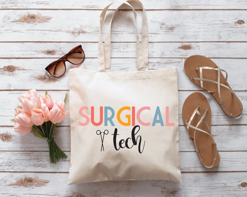 May include: A white canvas tote bag with the text "SURGICAL tech" in colorful letters and a pair of scissors graphic. The bag is on a white wood background.