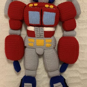 May include: A crocheted Optimus Prime toy with red, blue, and gray yarn. The toy is in robot mode with its arms and legs extended.