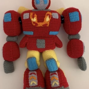 May include: A crocheted toy robot with red, yellow, blue, and gray accents. The robot has a large head with a blue face and a red helmet. The robot has large arms and legs.